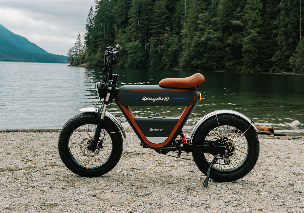 Side profile of the Rictor K1 electric scrambler e-bike, featuring a black frame with orange accents and a tan saddle, parked on a gravelly lakeside shore