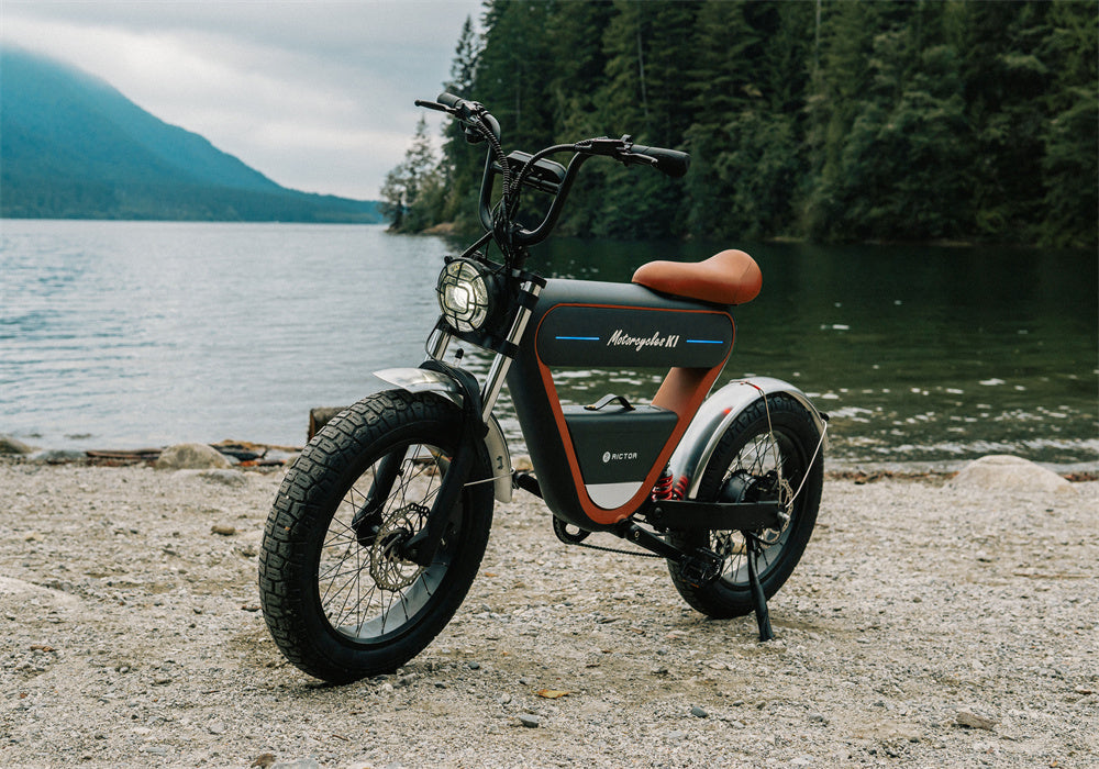 Rictor K1 moped-style e-bike in black with orange accents, parked on a gravel shore with a lake and forested hills in the background.