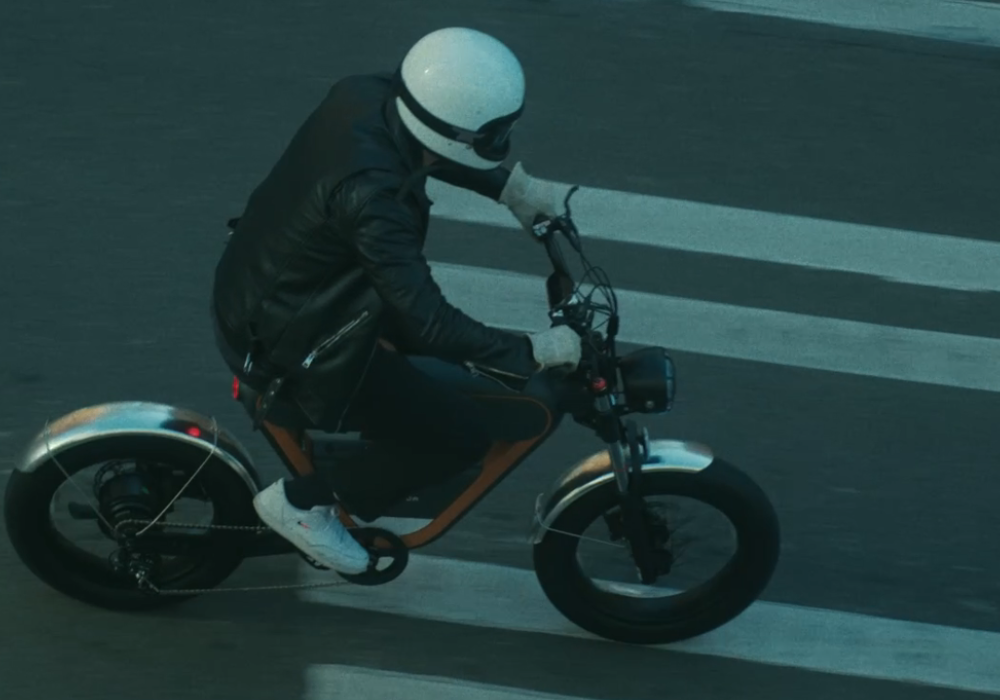 Rider wearing a white helmet and black jacket cruising on a RICTOR K1 cafe racer-style e-bike across an urban crosswalk.